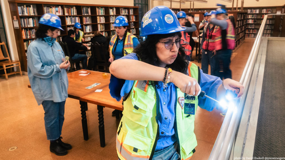 A person with long hair in a hard hat with a flashlight inspecting a railing in a library.
