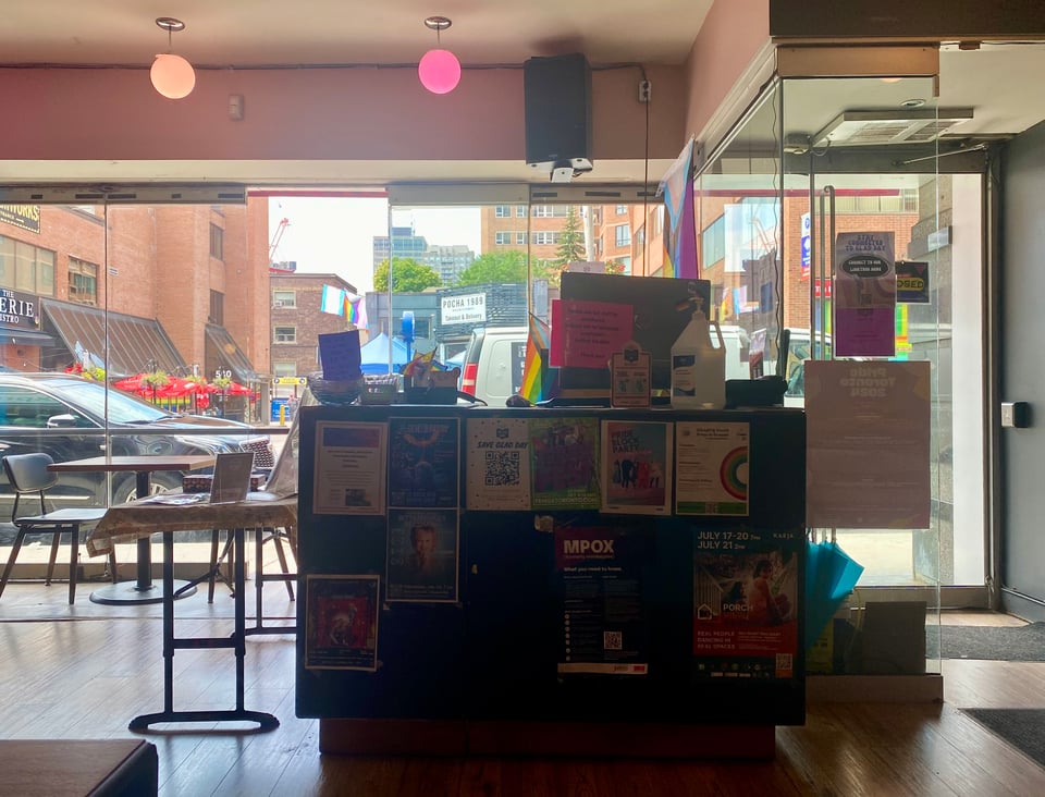 A view of the front desk of Glad Day Bookshop in Tkaronto, with the shop entrance to its left and floor to ceiling glass windows to its rear, offering local street views.