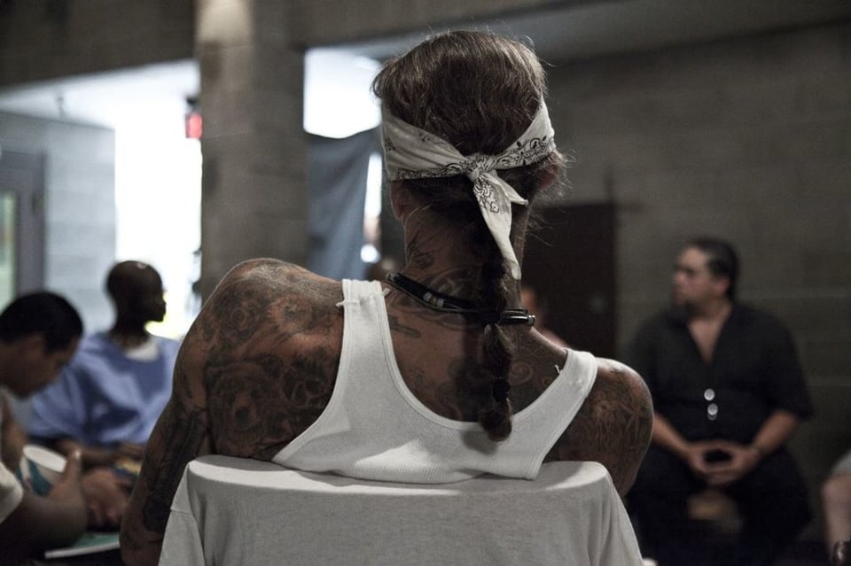 [In a film still of people sitting in a circle inside a cinderblock room, the camera focuses on the back of one of the members of the circle, who is wearing a white tank top and a white bandana around his head.]