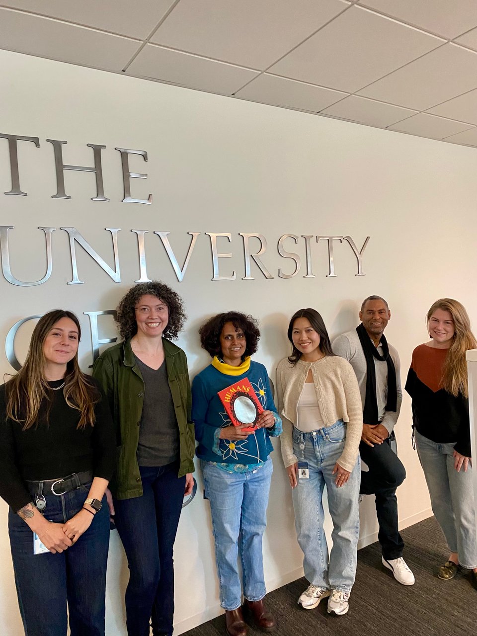 Five figures of various genders and ethnicities pose indoors in a relaxed fashion in front of a white wall with the University of California Press logo behind them in large letters. One of the central figures carries a bright red book with a large oval mirror and yellow lettering on it.