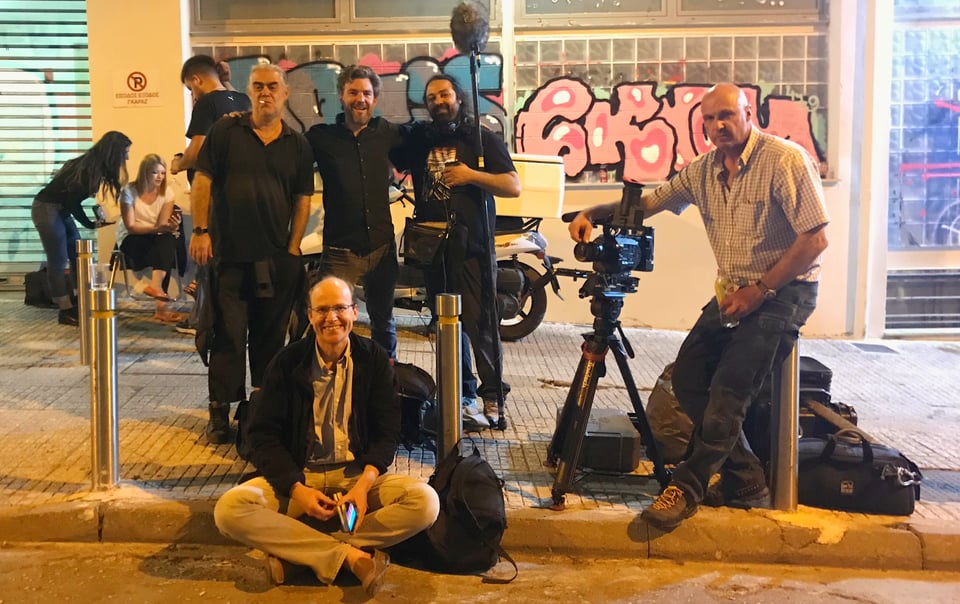 Photograph of a small film at the side of a street at night. There are 5 people, one of whom is resting his arm on a movie camera on a tripod, and one of who is holding a microphone boom.
