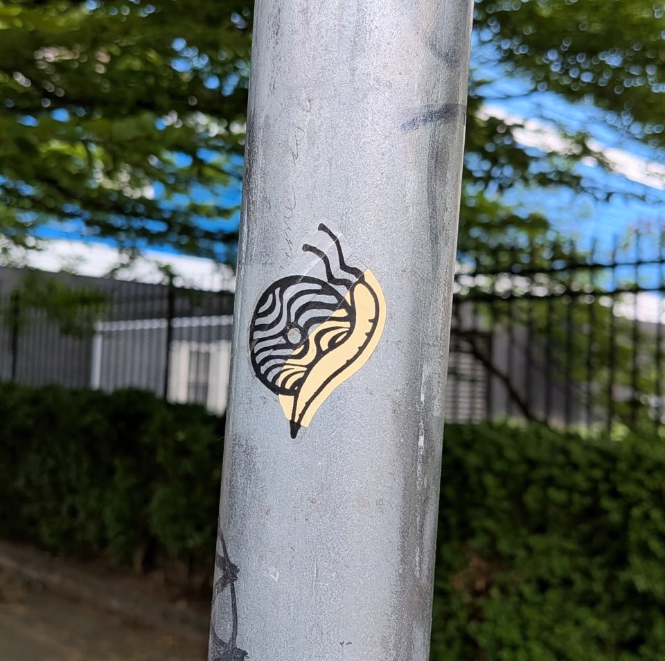 A close-up photo of a grey streetsign post with a black and yellow snail sticker crawling up it.