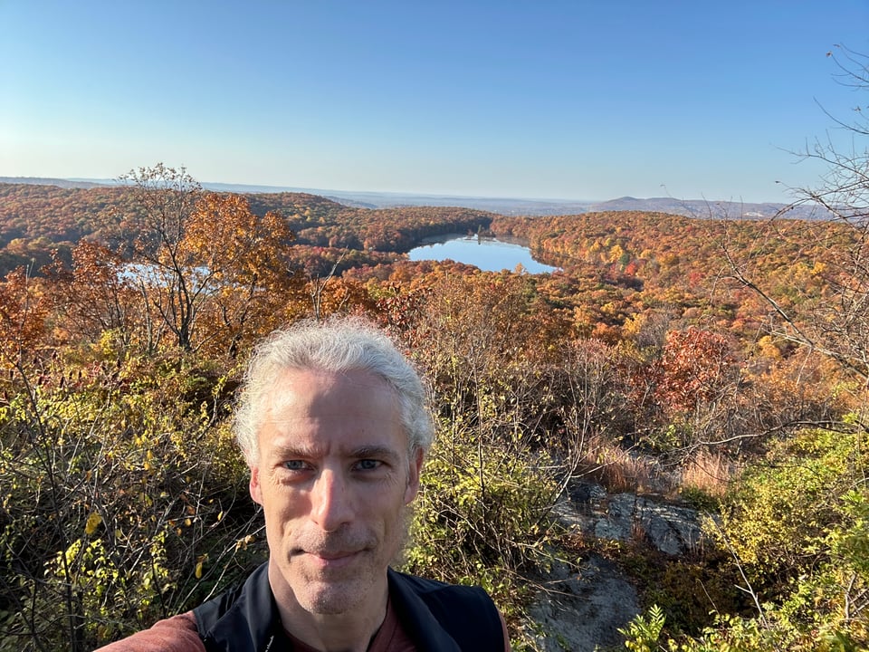 Selfie of white man standing above woods and a lake