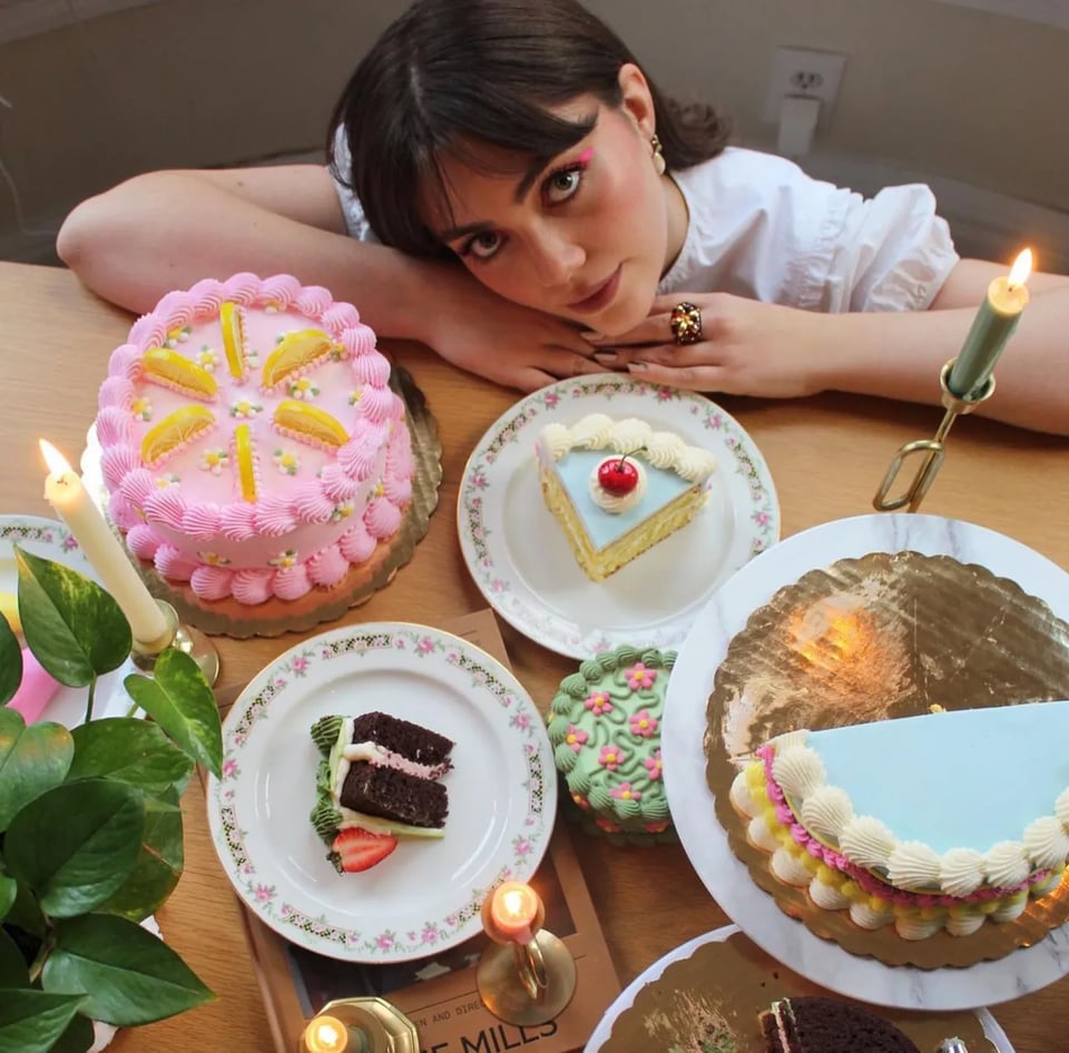 a picture of baker, Morgan Knight, resting on a table that holds several colourful cakes.