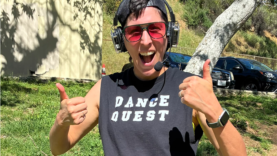 A woman wears a DJ headset with a black shirt saying Dance Quest gives two thumbs up.