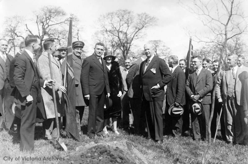 Black and white photo. A group of people standing around a pile of dirt and a tree. One person holds a shovel.