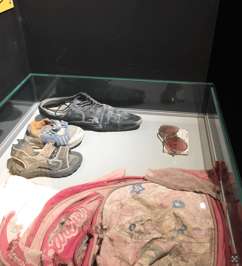 Photograph of a museum display case presenting worn shoes, red sunglasses, and a torn and dirty pink backpack with blue star decorations.