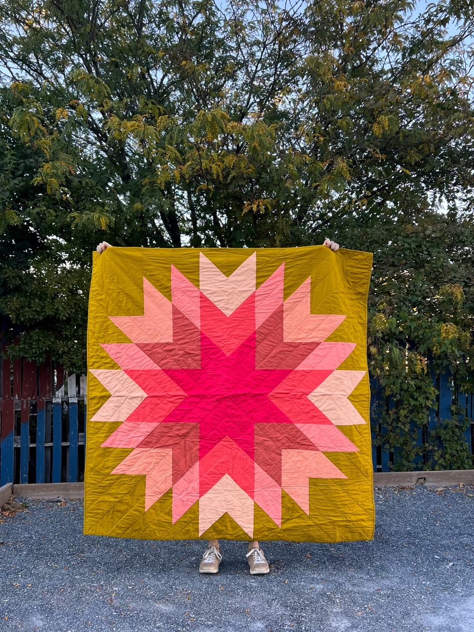 a star quilt in pinks and green being held in front of trees and a fence