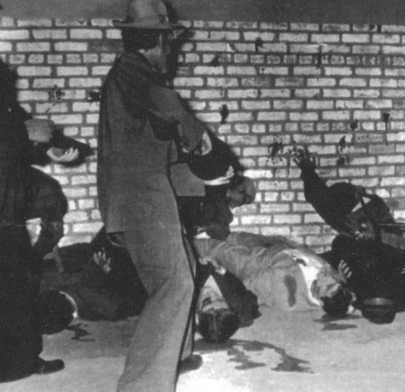 Black and white photograph that shows the reassembled wall with either people or mannequins (is not clear) posed as gunmen and victims