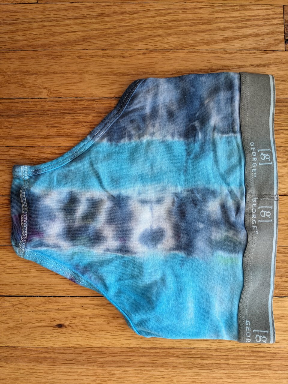 A pair of tie-dyed briefs, backside view with wide light and dark blue stripes and a gray waistband facing the side of the pic, against a hardwood floor.