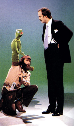 Jim Henson rests on one knee, reaching upwards with Kermit. Beside him, John Kleese looks down to make eye contact with the frog puppet.