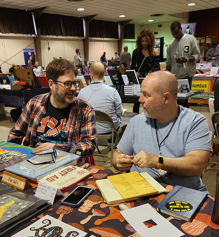 A picture of two white guys sitting behind a table at a small press convention. There are various books and pieces of artwork on the table.