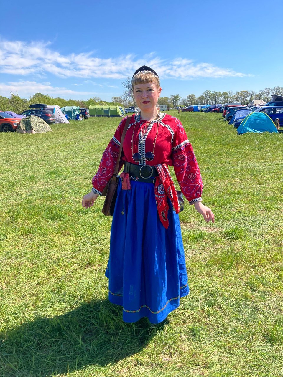 A middle aged white woman with blonde hair tied back with a fringe. She's standing on green grass in a field containing spaced out tents and cars. She's wearing a bright blue skirt, black leather belts and a bright red blouse. She has a pointed black headband in the eastern european style. Her clothing is adorned with embroidery and there are various pouches and trinkets strapped to her belt