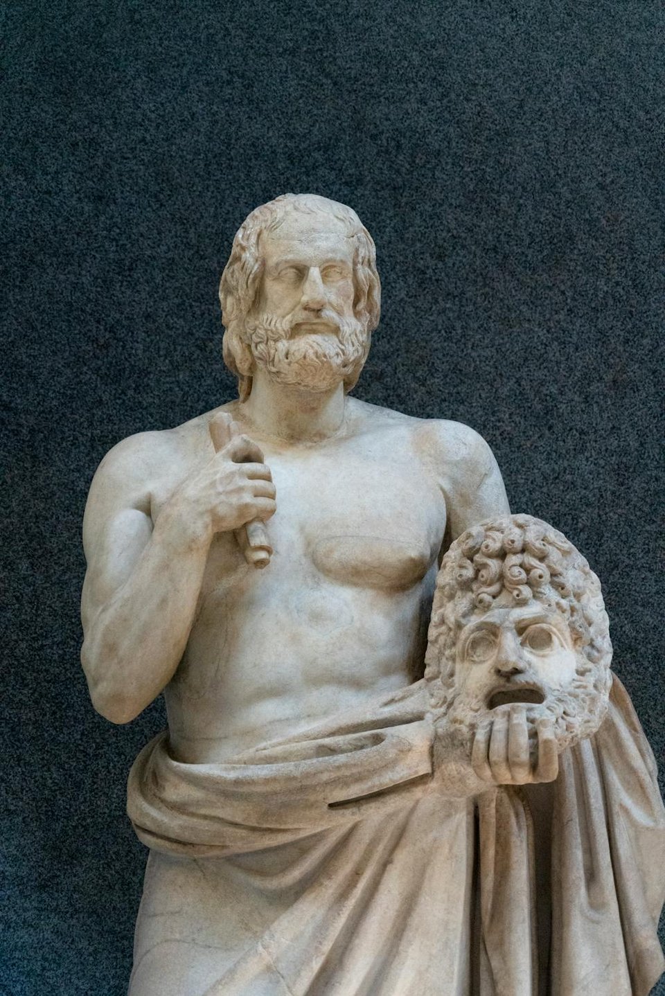 An ancient greek statue of a man holding a scroll in his right hand and a mask in his left; the mask has hollow eyes and an open mouth