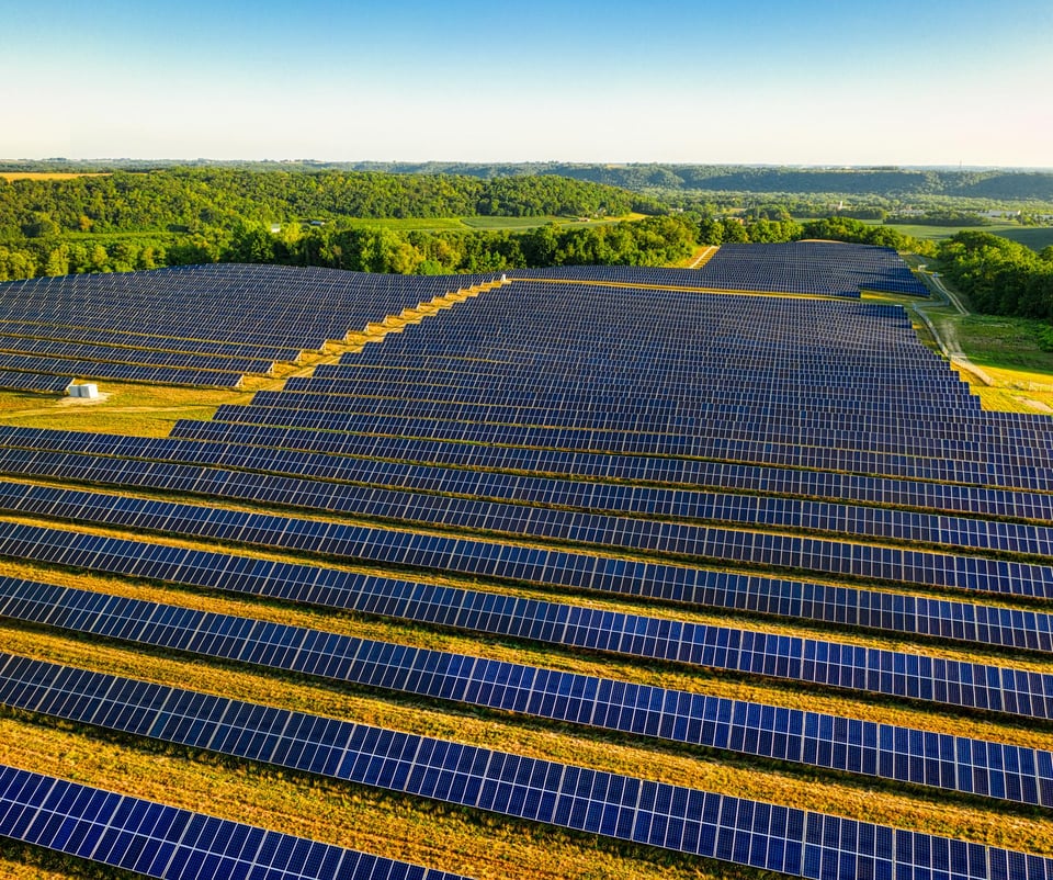 An aerial photo of a large solar farm with thousands of panels lined up in hundreds of rows. They are ringed by green treed forest land. The sky above is blue.
