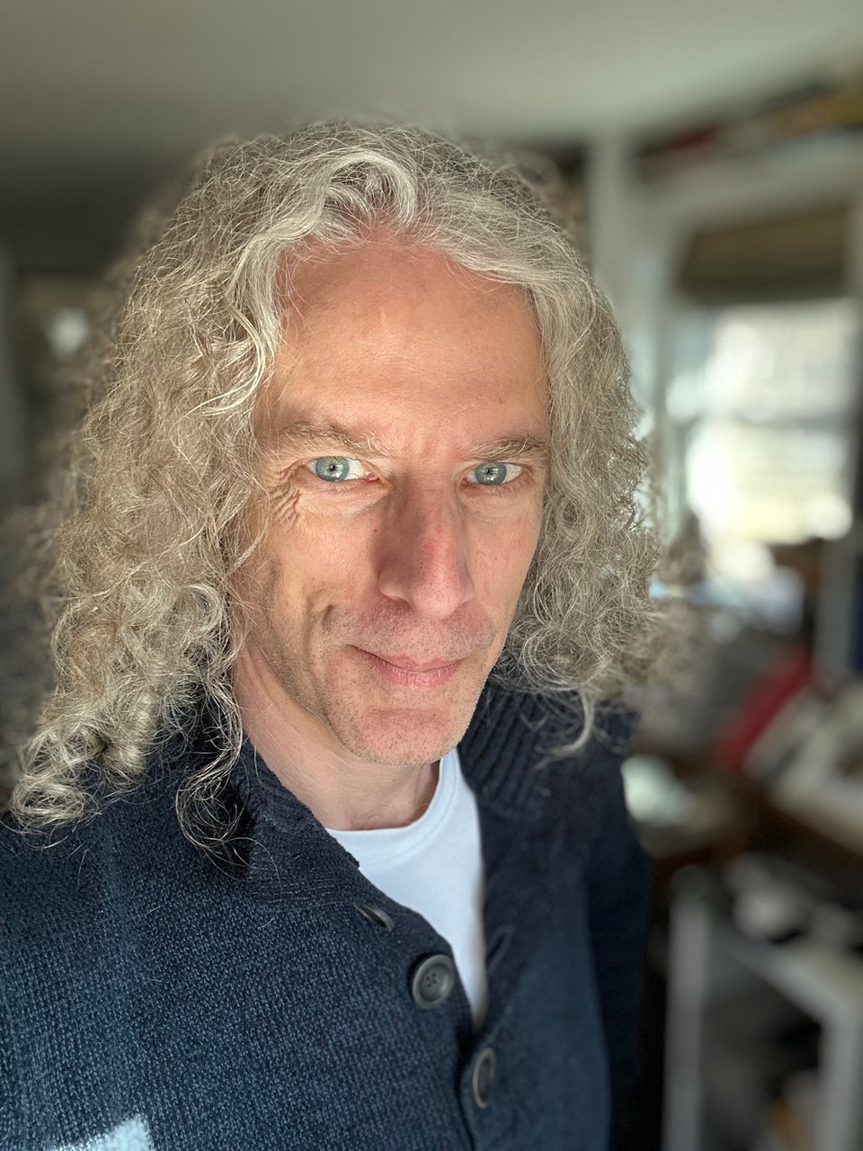 Selfie of white man in navy blue sweater; he has extremely poofy long curly hair, and the lighting makes his eyes look pretty bewitching