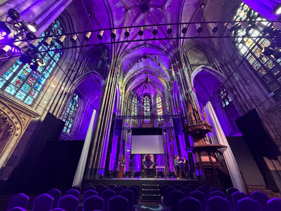 The interior of a gothic church lit in purple light with a stage by the altar and seats set up for the audience.