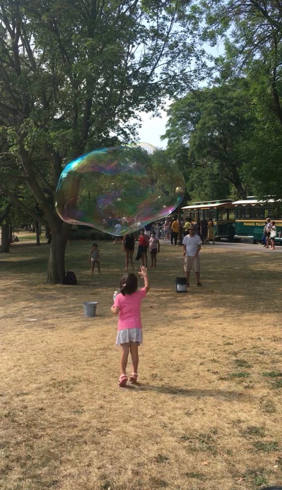 A little girl in a pink top and skirt reaches upward toward a huge, iridescent bubble that is bigger than her, floating above.