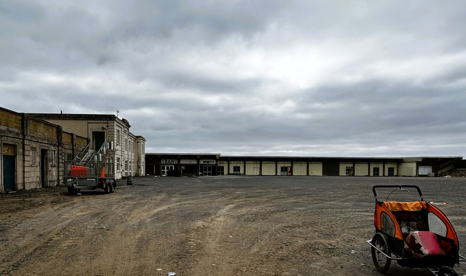 A derelict recreation ground within a 1930s lido. The rear of the changing block is on the left, and a single storey internal building is ahead, with a handpainted sign pointing to the bar. There's a cherry picker parked in one corner and, rather more randomly, an old bike trailer for kids on the other side.