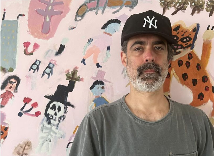 a bearded man posing in front of a painting featuring cartoon-like characters on a pink background
