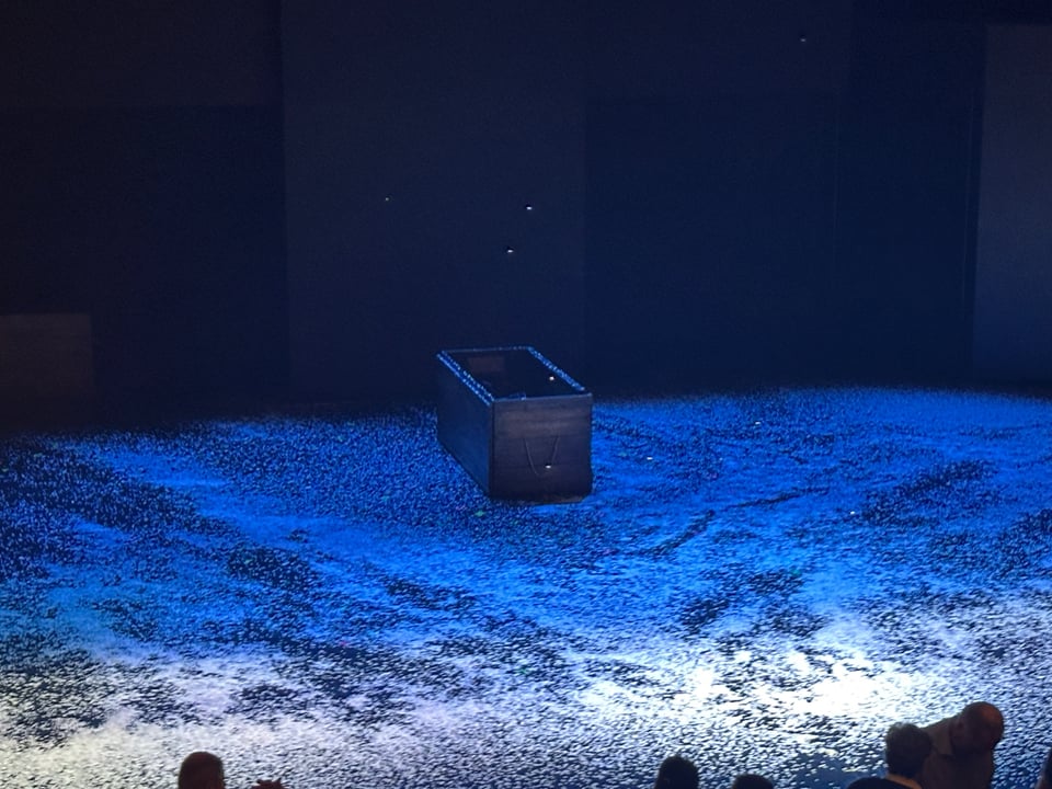 An empty coffin rests on the floor of a stage, which itself is dusted in snow.