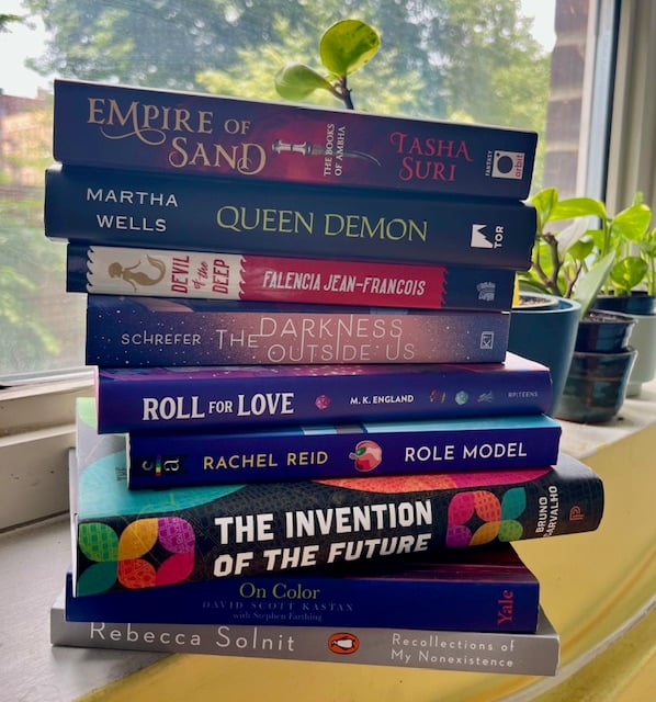 A precariously stacked tower of nine books on a windowsill, with green plants behind them and trees out the window.