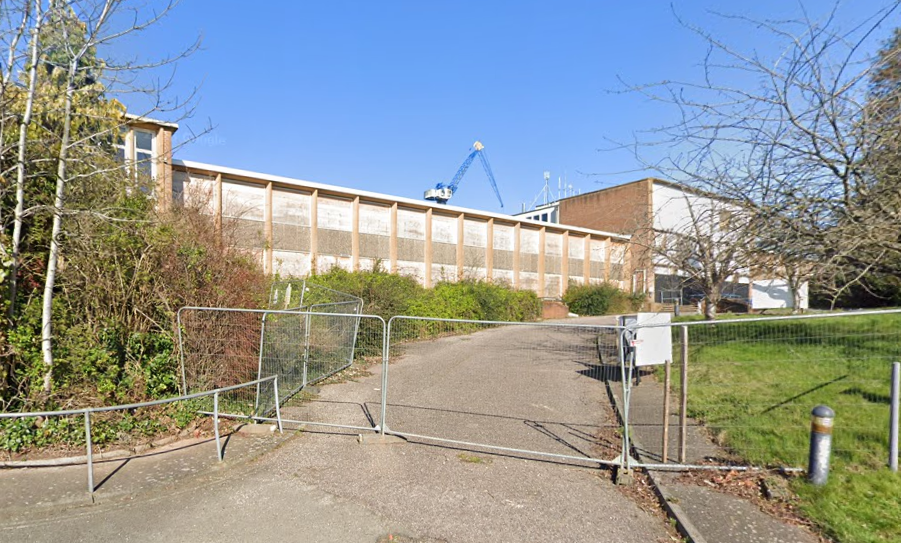 A boarded up 1950s building. It's mostly two storeys of concrete panel building, but rises to three stories and brick in a couple of places. The access road is fenced off.