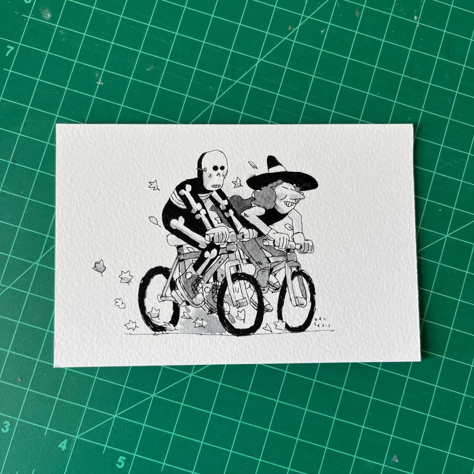 Two kids in halloween costumes, one a skeleton and one a witch, riding their bikes. Some fallen leaves are getting kicked up as they ride past.