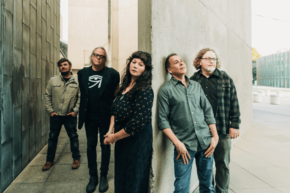 Two men and a woman stand in a narrow space between two walls, while on the other side of the wall to the woman's left stand two more men.