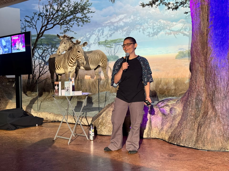 Photo of me (Danna Staaf) giving a talk in African Hall in the California Academy of Sciences. I'm smiling and holding a microphone in front of a display that includes two zebras. On a screen off to one side you can see pictures of a cuttlefish and a sea hare both releasing ink.