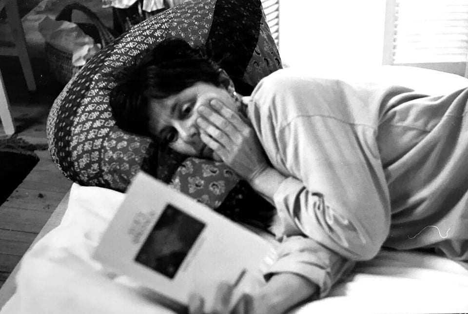 Helen garner lying down in bed, left hand pressed to her face, right hand holding open a book