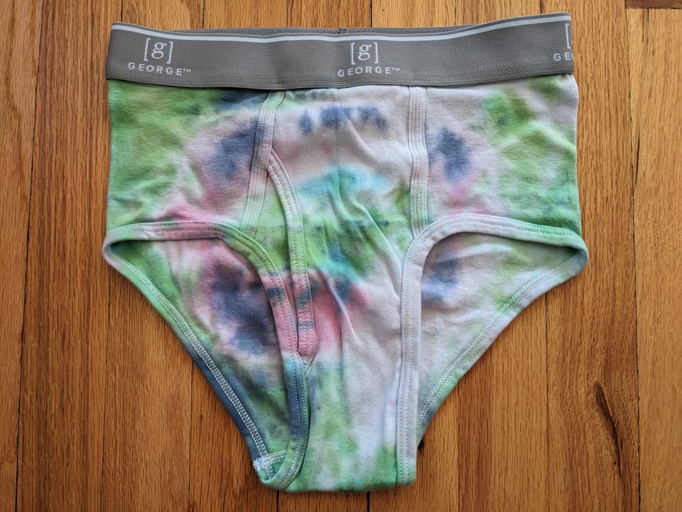 A pair of tie-dyed briefs, green and blue and pink in a very light bullseye shape, with a gray waistband, against a hardwood floor.