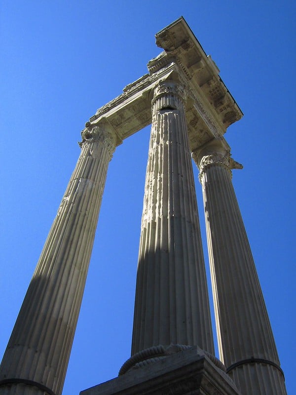 A low-angle view of three tall, fluted Roman columns with ornate Corinthian capitals, supporting a partially intact entablature against a vivid blue sky.
