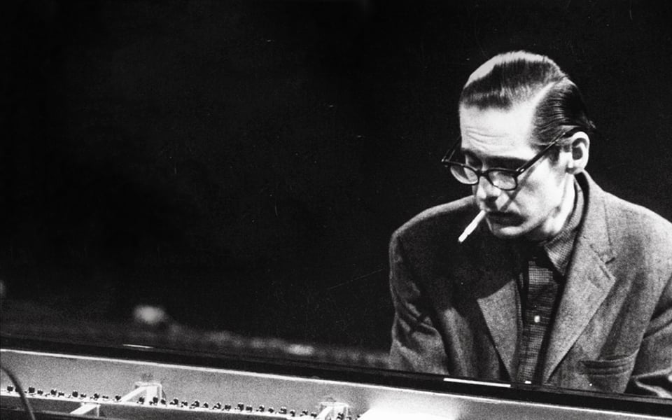 A white man sat at a piano. He has a cigarette in his mouth and he is wearing black rimmed glasses.