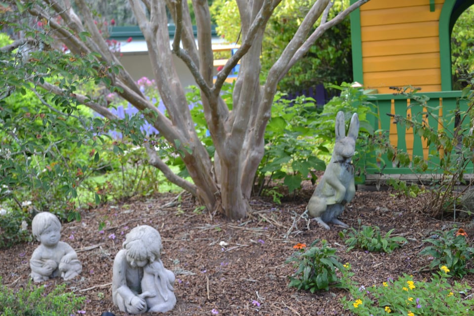 Two sculptures of babies and one of a really creepy bunny in a small flower bed with a green and yellow building behind.