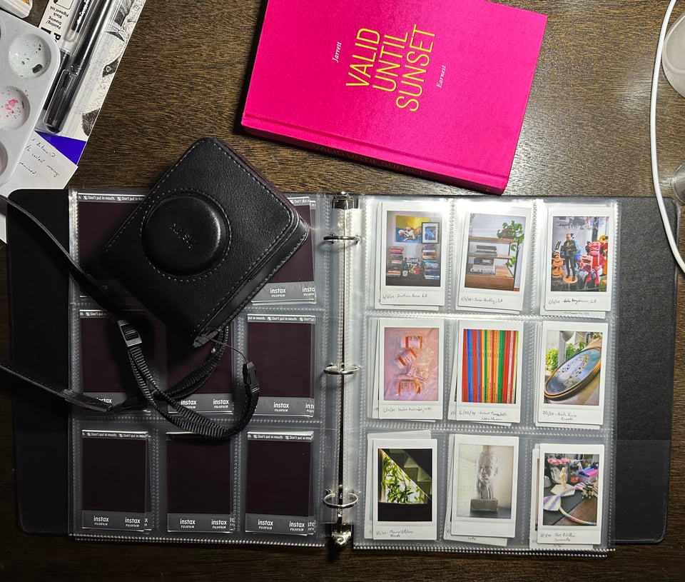 Photo of a drawing table that contains a Fuji Mini-Evo camera in its case, a copy of the art book VALID UNTIL SUNSET by Jarret Earnest, and a black binder opened to a page of Instax photos in a plastic sleeve