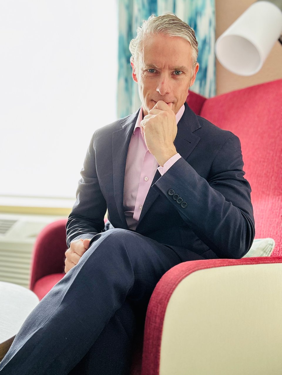 Photo of white man in a navy suit & pink dress shirt, sitting in a red chair, with one leg over the other knee