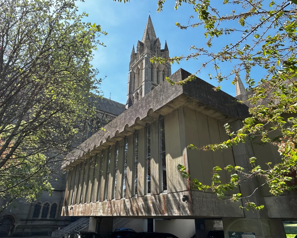 In the foreground is the cast concrete building. From this angle, you can see the rough cast to the concrete, and the semicircular detail to the window slits. Behind it is the very tall, very pointy spire. All around there are trees coming into leaf.