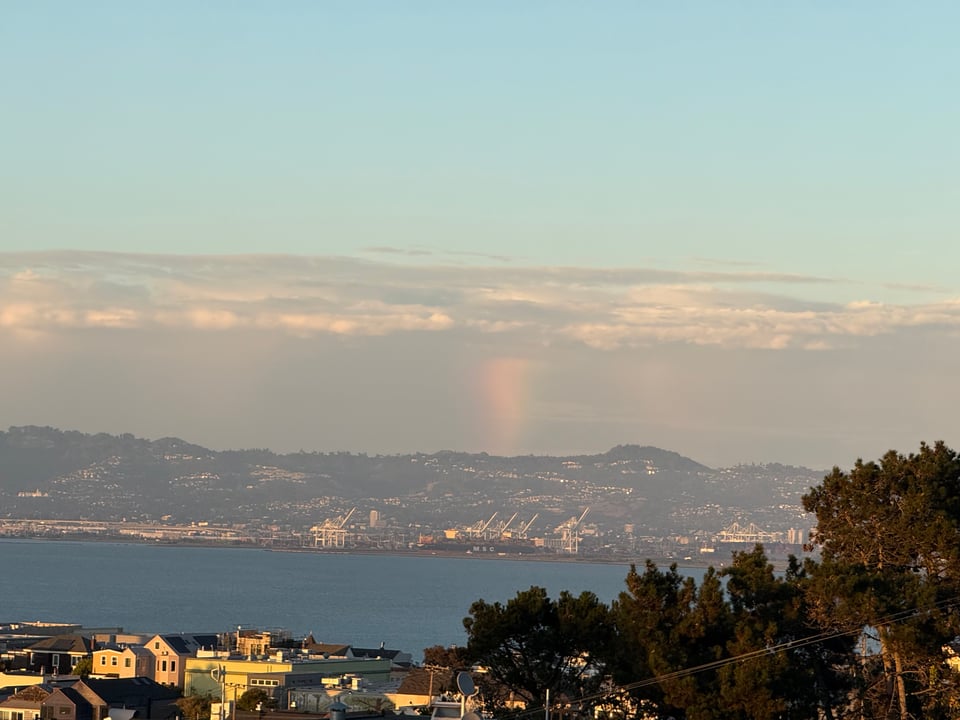 A distant view of a hazy landscape featuring a harbor and mountains under a soft sky, with a faint rainbow visible among the clouds.