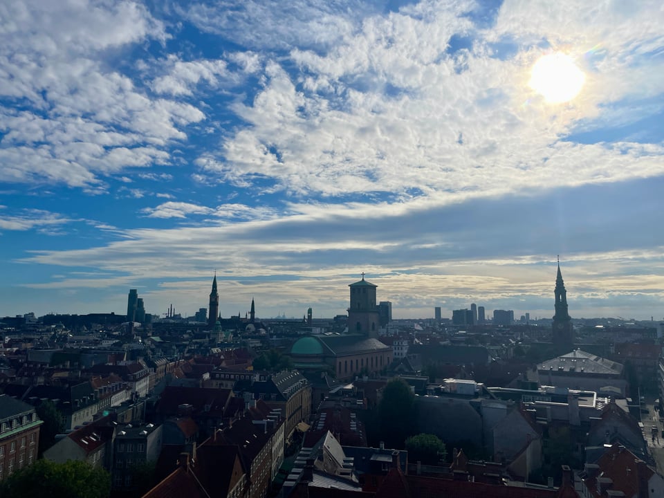 City skyline of Copenhagen with half a dozen towers and spires in the distance. The blue sky is loosely covered with clouds and the sun shines on the top right of the frame.