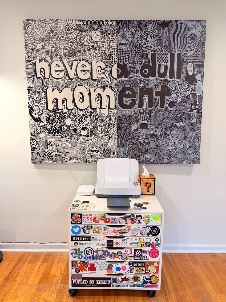 A large black and white intricate line drawing painting with text reading "never a dull moment". Beneath it is an Ikea storage cart covered with sticker labels, supporting a printer and tissue box.