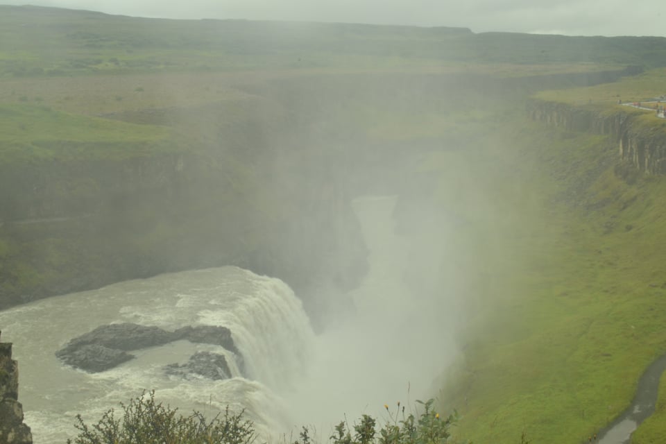 A gorge with green grass on either side. A big waterfall falls off the side of the gorge, sending up a spray of mist.