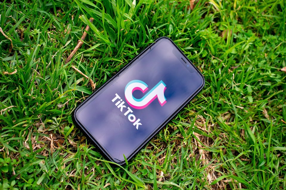 An Iphone lying in a field of grass, with the Tik Tok logo on its otherwise blank, black screen. It is seen from above