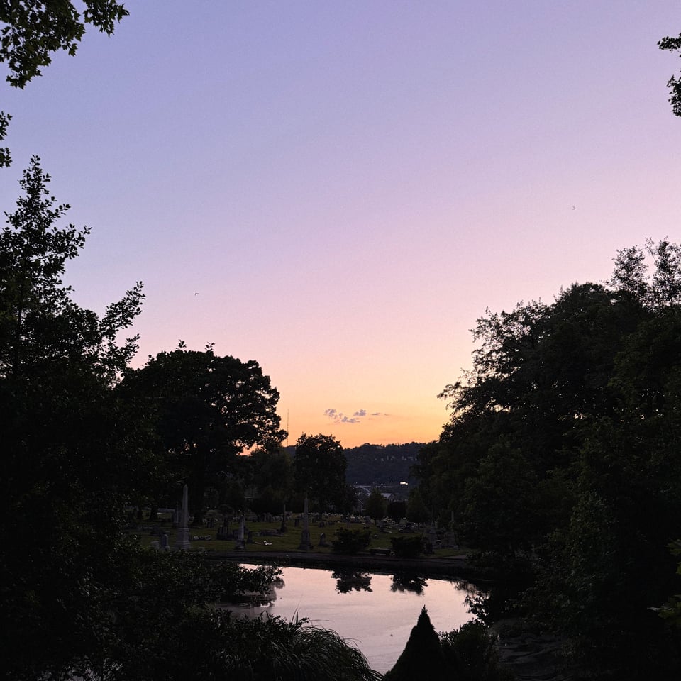 the very edge of a sunset in a graveyard, summertime, sky and trees reflecting off of a small pond in the dusk