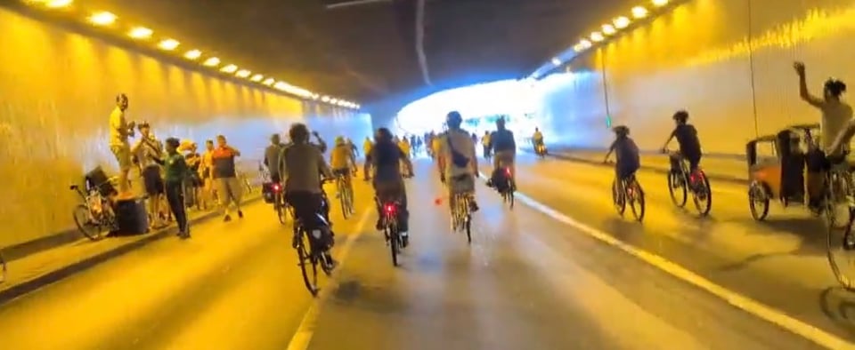 Cyclists going through a tunnel with dancers and speakers along the side.