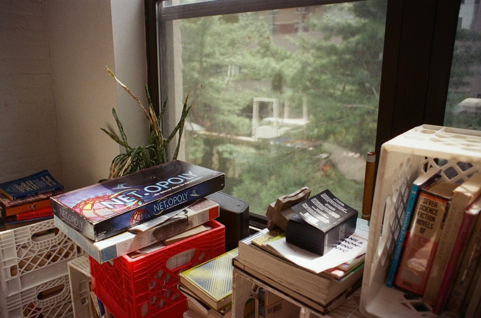 [Milk crates in front of a window hold stacks of books. Topping one of the stacks are two board games, the visible one reads Net-opoly.]