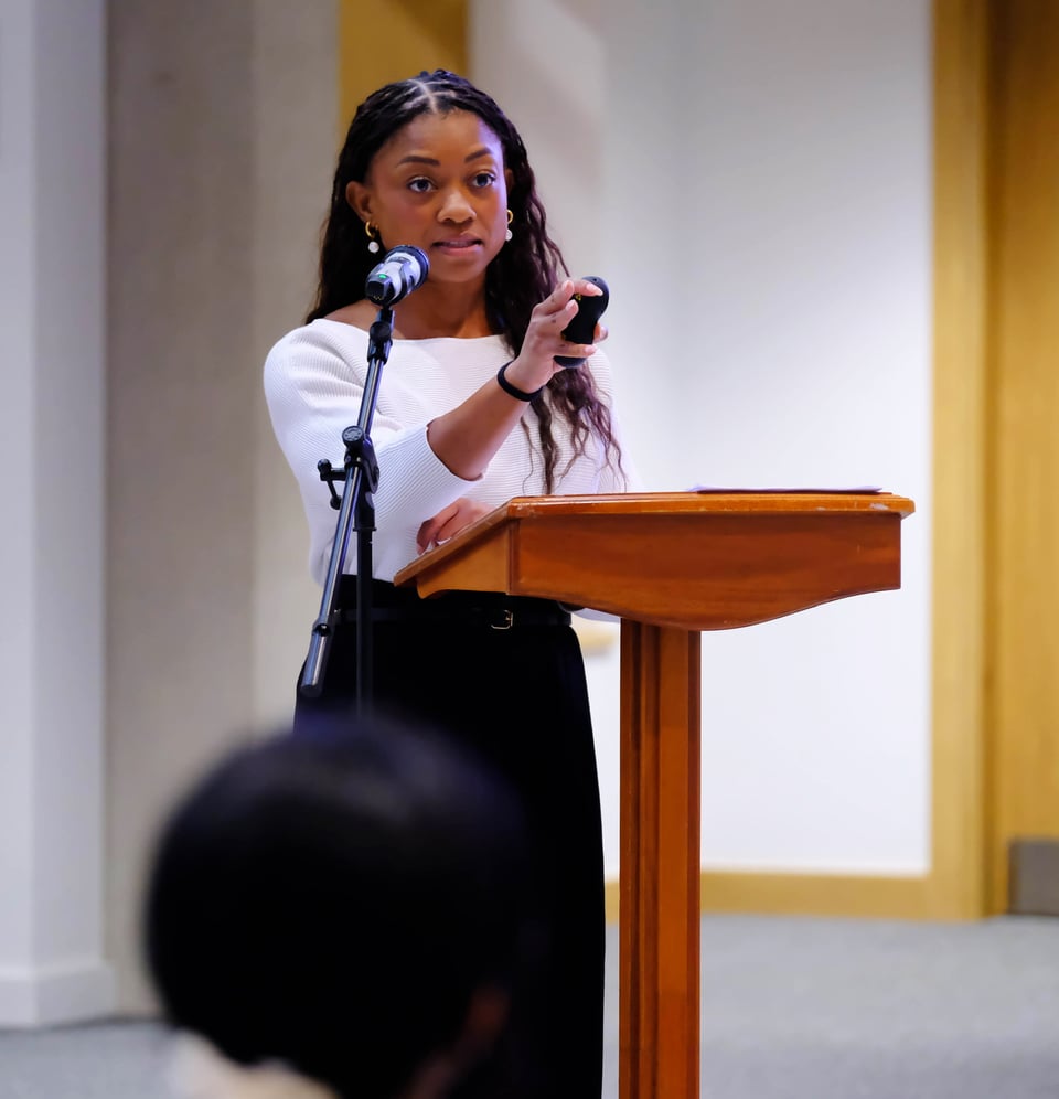 Nadia stands behind a wooden lectern, gesturing towards the audience with a small black presentation remote. She wears a white top and black trousers, with a microphone positioned in front of her.