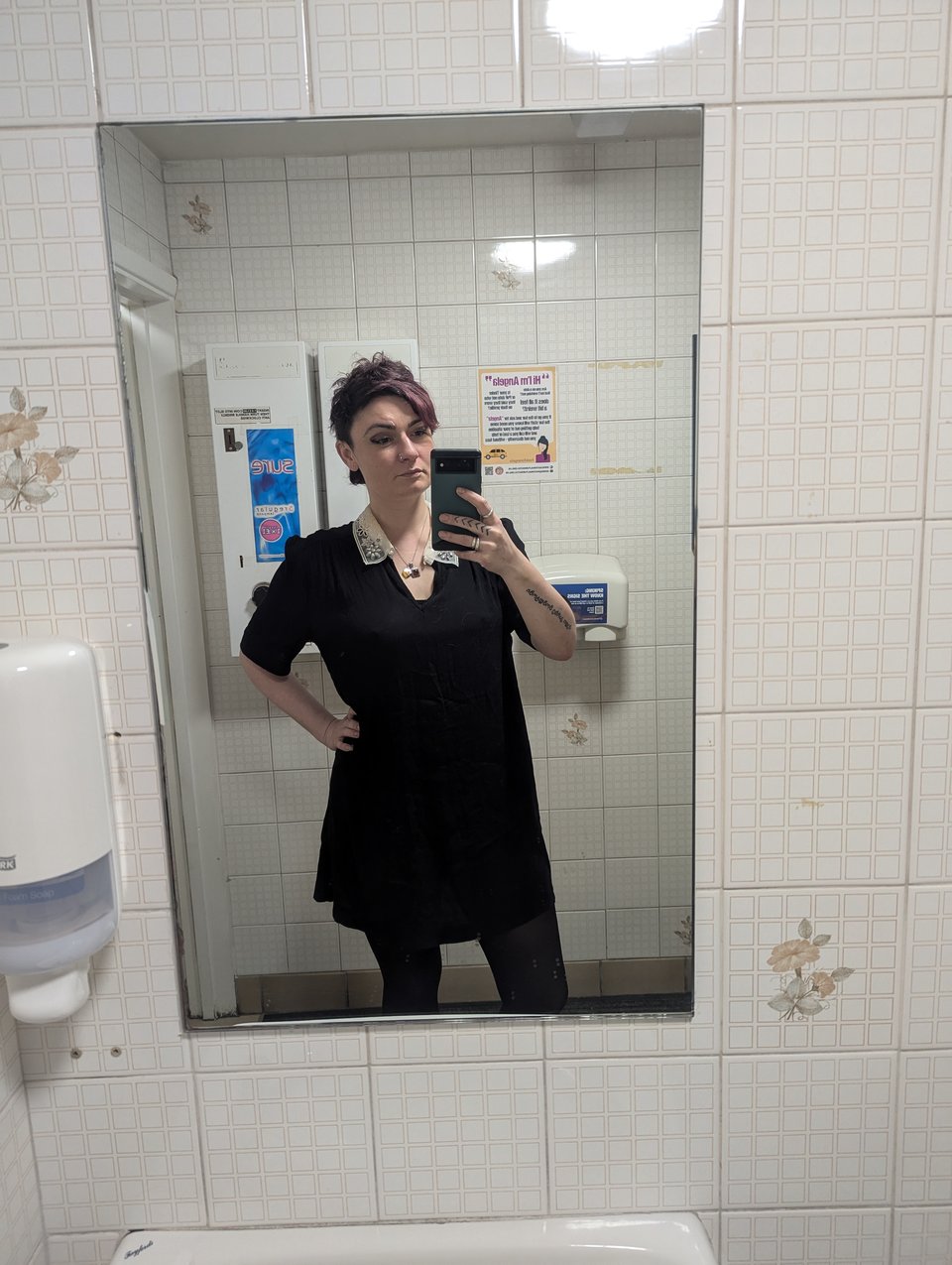 A woman with short hair wearing a black dress with a white collar in an unglamourous pub toilet