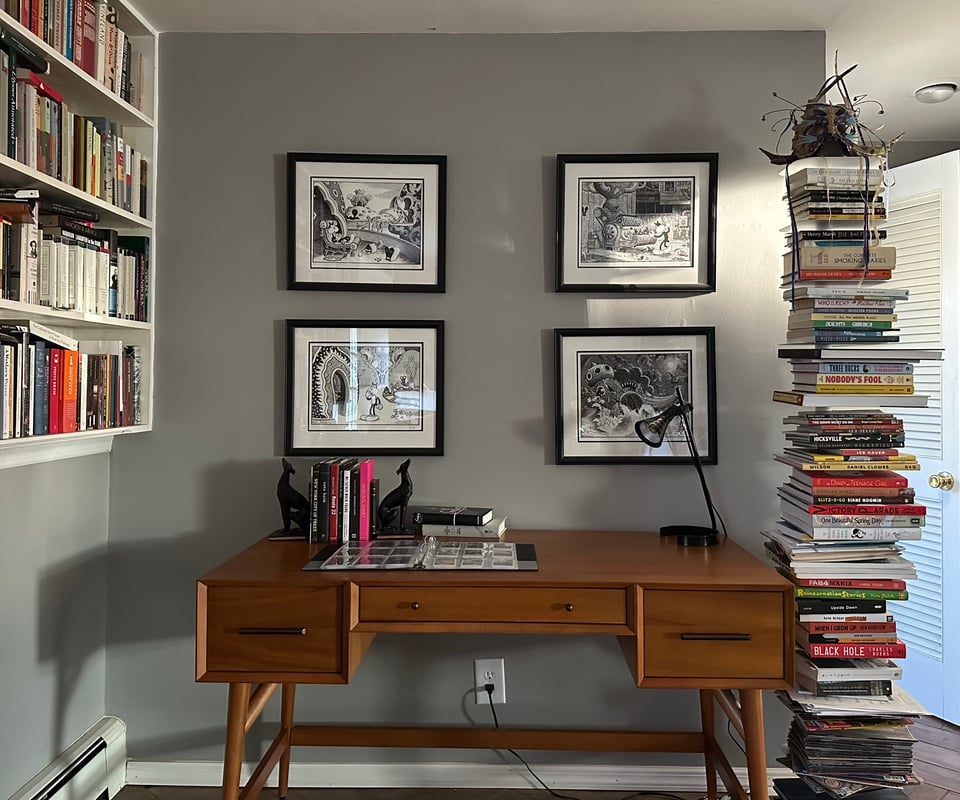 photo of an acorn-colored midcentury desk with a pair of greyhound bookends on it holding a few books; on the gray wall behind the desk are 4 framed prints by Jim Woodring; to the right of the desk is a vertical bookshelf loaded with books and comics; to the left is a partial view of built-in bookshelves along a wall