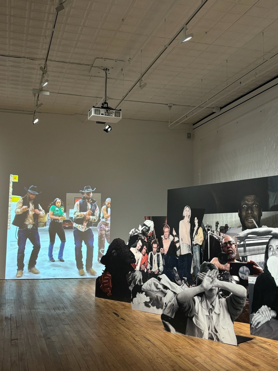 [A multimedia installation with a video of people dancing in cowboy hats in the background, and big blow-ups of photographs and cardboard cut-outs assembled on the floor in the foreground.] 